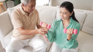 Yaşlı kadın oturma odasında dengeyi ve koordinasyonu arttırmak ya da ortak sağlık ve esnekliği artırmak için dambıl tutarken mutlu büyükbaba büyükanneyi cesaretlendiriyor ve destekliyor. Myrmidon.