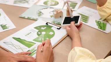 İş ekibi, temiz enerjiyle temsil edilen yel değirmeni modeline odaklandı. İş arkadaşı, karbonu azaltmanın yolunu seçen yeşil güç grafiğini analiz ediyor. Ahşap blok sembolize ekoloji evi masaya koyuldu. Tanımlama.