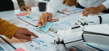 Çağdaş bir ofis ortamında bir insan eli ve bir robot kol işbirliği yapar. Sahne, yeniliği sembolize eden grafikler ve veri analizi ile etkileşimi vurgular. Noogenesis