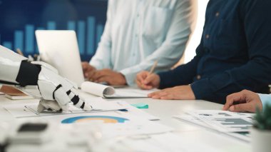 Bir insan takımı modern bir ofiste bir robotla işbirliği yapıyor, finansal tabloları analiz ediyor ve verileri tartışıyor. Bu sahne iş dünyasındaki teknoloji ve takım çalışmasının kaynaşmasını vurgular. Noogenesis