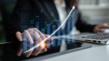 Modern ofis ortamında dijital grafikleri ve eğilimleri analiz etmek için tablet kullanan bir iş adamı. Teknolojiyi ve veriye dayalı karar verme mekanizmasını vurguluyor. Etki