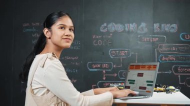 Akıllı kız karatahtadaki dizüstü bilgisayar kodlama ya da programlama sistemini kullanıyor. Bilgisayarda çalışan mutlu bir lise öğrencisi minyatür kalp gösteriyor ve BTMM sınıfında kameraya bakıyor. Öğretim