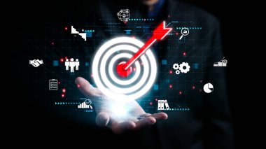 Bir iş adamı, analitik, büyüme ve başarıya odaklı stratejileri temsil eden dijital simgelerle çevrili bir el hareketiyle hedeflere ulaşma kavramını sergiliyor. Dörtgen