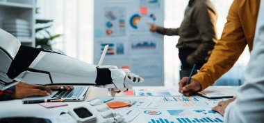 İşbirlikçi veri analizinde yardımcı bir robot eli gösteren fütüristik bir çalışma alanı. Profesyoneller modern takım çalışmalarının dinamiklerini ön plana çıkaran çizelgeler ve teknolojiyle ilgilenirler.