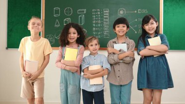 Çok kültürlü bir ilkokul öğrencisi, günlük kıyafetleriyle sınıfta dikilirken kameraya bakıyor. Bir grup tatlı çocuk ellerinde defterle sınıftaki tahtada kameraya gülümsüyor. Pedagoji.