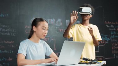 Genç ve güzel akademik öğrenci yazma, kodlama ve programlama sistemini bilgisayarı ya da aygıtı olan masaya yerleştirirken Afrikalı çocuk metaevrene girmek için kulaklık takıyor. yenilikçi eğitim. Öğretim