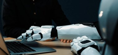 İnsan-robot etkileşiminin yakın plan görüntüsü, masadaki dizüstü bilgisayara uzanan bir robot kolu gösteriyor, teknolojinin ve işbirliğinin geleceğini yansıtıyor. Noogenesis
