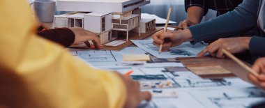 Canlı bir çalışma alanı, mimari tasarımlar üzerinde işbirliği yapan, çizim yapan, planları inceleyen ve modern bir proje için ölçek modelini inceleyen bir ekibi sergiliyor. SACTR