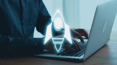 Modern bir çalışma alanında dizüstü bilgisayarda çalışan bir adam, dijital bir roket sembolü ile yenilik ve büyüme temalarını vurguluyor, iş stratejilerini ve gelecekteki olasılıkları yansıtıyor. Dörtgen