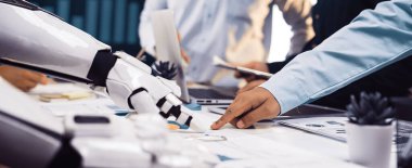 Bir insan eli ve robot kolun yer aldığı ortak bir sahne. Veri tabloları ve grafiklerle bir toplantıya katılıyor, iş planlama teknolojisinin bütünleşmesini vurguluyor. Noogenesis