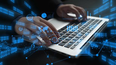 Arka planda canlı veri görselleştirmeleri olan bir dizüstü bilgisayarda yazan bir kişi, teknoloji, iş analizleri ve yenilikleri sembolize ediyor. Xenic