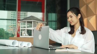 Genç ve güzel mimar mühendisin çatı malzemelerini kontrol etmesi ve ofisteki masaya yerleştirilmiş proje planı ve ev modeliyle dizüstü bilgisayarı kullanması. Yaratıcı iş tasarımı konsepti. Yönlendirici.