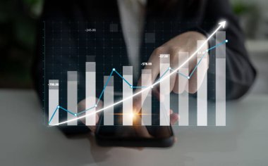 Bir iş adamı finansal verileri analiz etmek için akıllı telefon kullanır, grafik ve grafiklerle büyümeyi gösterir, modern finansal yönetim tekniklerini temsil eder. Etki