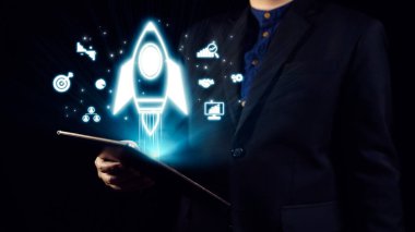 Bir iş adamı, teknolojideki yenilik ve büyümeyi ve başlangıç girişimlerini sembolize eden parıldayan bir roket ikonu ve çeşitli dijital sembollerle bir tablet tutuyor. Dörtgen