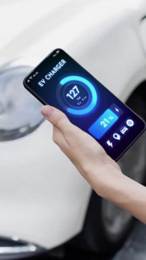 Akıllı telefon akü durumu arayüzünü elinde tutan iş kadını akıllı EV mobil uygulama ile otomobil parkındaki şarj istasyonundan elektrik şarj eden çevre dostu EV araba. İnceleyin