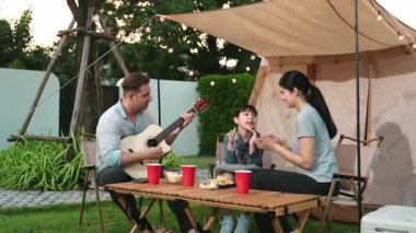 Baba gitar çalarken anne ve kız birlikte kampta oturuyorlar. Ebeveynler, genç nesil çapraz kuşak farklılıklarıyla zaman geçirmek için açık hava kamp aktivitelerini kullanıyorlar. Uyumsuzluk.