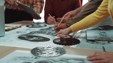 Bir grup mühendis, teknik çizimler ve araçlarla dolu bir masanın etrafında toplanarak, modern ofis ortamında işbirlikçi bir beyin fırtınası yapıyorlar. SACTR