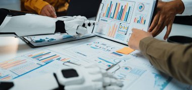 Canlı grafikler ve çizelgeler ile kompleks veri raporlarını analiz etmek için birlikte çalışan insan ve robotik elleri resmeden modern bir ofis sahnesi, işbirliği ve yenilikleri vurguluyor.