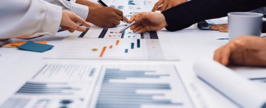 Katılımcılar, masadaki ayrıntılı çizelge ve grafikleri analiz ederek, modern bir ofis ortamında takım çalışması sergileyerek, işbirlikçi bir iş toplantısına giriyorlar. SACTR