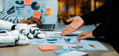 Etkili karar verme işlemleri için dizüstü bilgisayarlar ve çizelgeler ile veri analizi ve iş stratejisine odaklanan, insan ve robot arasındaki modern bir çalışma alanı gösterimi.