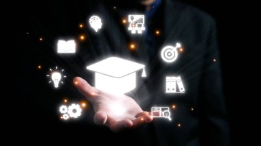 Çeşitli eğitim simgeleriyle çevrili bir mezuniyet şapkası sembolüne uzanan bir el, karanlık bir arkaplanda bilgi, gelişme ve akademik başarıyı sembolize ediyor. Dörtgen
