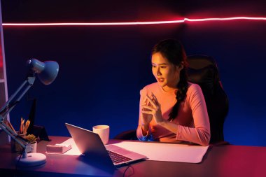 Geceleri sosyal medyada canlı yayında pozitif podcastla konuşan gülümseyen genç, güzel Asyalı etkileyici pembe neon ışıklı modern stüdyoda sunum için dizüstü bilgisayar kullanıyor. Taktik.