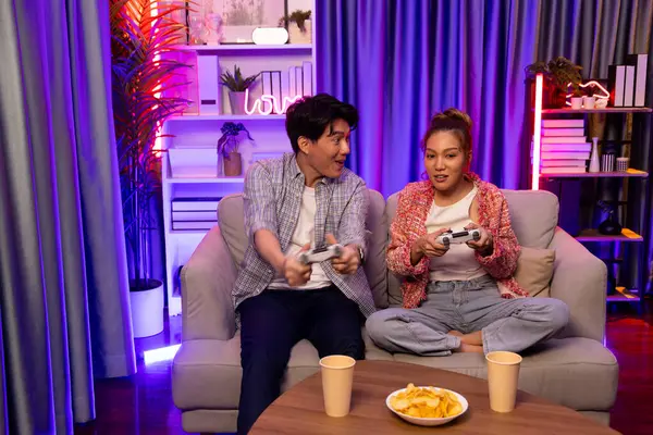 Neşeli, gülümseyen bir çiftin koltuğunda oturup oyun oynuyorlar. Oyun konsolu, hazır aperatifler ve içecekler, modern neon ışıklı, rahat oturma odasında eğlenceli bir rekabet seviyesinde. İnfobahn.