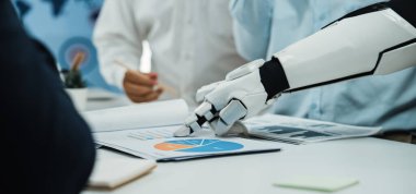 Robotik bir el modern bir ofiste yapılan ortak bir toplantıda teknolojinin kurumsal stratejiler ve analizlere entegrasyonunu gösteren bir iş grafiğine işaret eder.