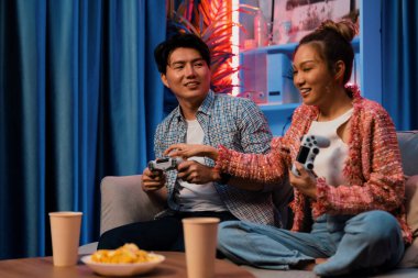 Neşeli, gülümseyen bir çiftin koltuğunda oturup oyun oynuyorlar. Oyun konsolu, hazır aperatifler ve içecekler, modern neon ışıklı, rahat oturma odasında eğlenceli bir rekabet seviyesinde. İnfobahn.