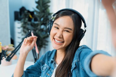 Sunucu kanalı genç ve güzel Asyalı kız akıllı telefonuyla selfie çekiyor. Modern stüdyo ofis konseptinde kulaklık takıyor. Sahne arkası yayıncısı sosyal medyada canlı yayın yapıyor. Taktik.