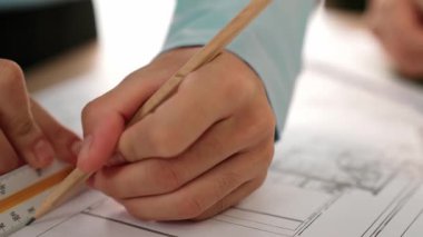 Mimarın bir videosu taslağı ölçmek için cetvel ve kalem kullanır. Mimar, mimari planı etrafa dağılmış bir şekilde stüdyoda bir masanın üzerine ev inşaatı tasarlıyor. Eline odaklan. Tanımlama.
