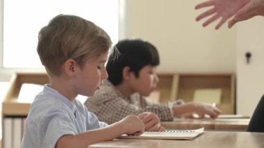 Beyaz tenli ilkokul öğrencisinin tahtada otururken bir nota bakması ve soruya cevap vermesi. Çocuklar derse katılırken genç öğretmen sınıf çalışmasını ya da sınavı açıklar. Pedagoji.