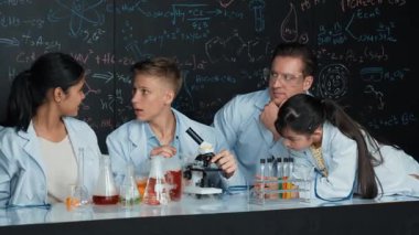 Çok kültürlü bir öğrenci mikroskop altında incelerken güzel bir kız da numuneyi inceliyor. BTMM sınıfında karatahta bilim teorisi deneyi yapan liseli çocuklara bakan öğretmen. Öğretim