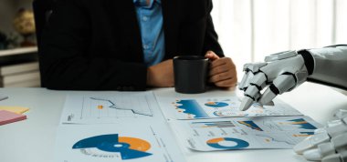 Bir iş adamı modern bir masada oturur, çizelge ve grafikleri robot koluyla analiz eder, teknolojiyle insan öngörüsünü gelecekteki ofis ortamında birleştirir. Noogenesis