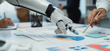 Robotik bir el modern ofis ortamında iş grafiklerine uzanıyor, çeşitli profesyoneller arasında bilgilendirilmiş kararlar için verileri analiz eden bir işbirliği sergiliyor.