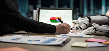 Bir insan eli kağıda çizerken bir robot el yardımcı oluyor, iş analizlerinde verileri görselleştirme ve üretkenliğe odaklanmış modern bir ofis ortamı sergiliyor. Noogenesis