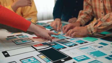 Çağdaş bir çalışma alanında renkli prototipler ve çizimlerle etkileşim içinde olan akıllı telefon uygulama tasarımı ile uğraşan bir ekibin canlı bir sahnesi. SACTR