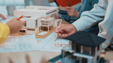 Bu resim, ellerin mimari model tasarımıyla meşgul olduğu, planlar ve 3 boyutlu ev modelleriyle çevrili, yaratıcılığı ve takım çalışmasını ön plana çıkaran bir çalışma alanını gösteriyor. SACTR