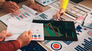 Modern bir iş ortamında takım çalışmalarının ve stratejik tartışmaların vurgulandığı, veri analizi ve grafiklerle uğraşan dinamik bir işyeri sahnesi. SACTR