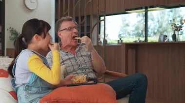 Grandfather and granddaughter eat french fries watch interesting entertainment on TV. Senior use technology communicate with young generation cross generation gap strengthen family bond. Divergence.