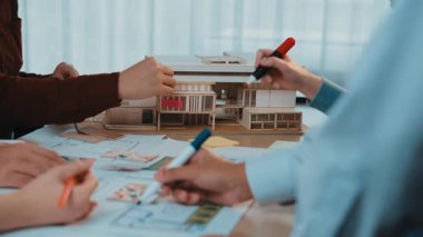This image captures a vibrant architecture planning session with hands engaged in sketching designs and assembling a miniature model in a modern design studio. SACTR
