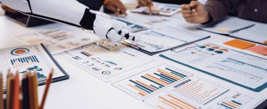 Robot bir kol ve insan elinin yer aldığı modern bir iş toplantısı sahnesi. Çeşitli çizelge ve raporları analiz etmek, takım çalışmasını ve teknoloji entegrasyonunu resmetmek. Noogenesis