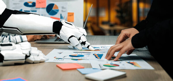A robotic arm collaborates with a human in analyzing business reports and data visualizations in a modern office setting, enhancing productivity and innovation.Noogenesis