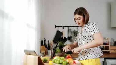 Kafkasyalı anne, modern mutfakta dizüstü bilgisayarda yemek tarifine bakarken salata yapmak için yemek hazırlıyor. Yetenekli ev bekçisi taze sebze kullanarak kahvaltı hazırlıyor. Pedagoji.