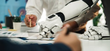 Robotik bir el ve insan eli, modern çalışma alanlarındaki otomasyon ve takım çalışmasını sembolize eden gösterişli bir ofis alanında proje planlamasında işbirliği yapmaktadır.