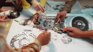 A focused group engages in a technical discussion at an office desk, analyzing mechanical designs and diagrams while holding tools and documents, showcasing teamwork. SACTR
