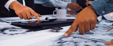 Dinamik bir iş toplantısı sahnesinde, dijital cihazların yanı sıra anahtar ayrıntıları da vurgulamak için elleri kullanarak masada teknik planları tartışan meşgul profesyoneller yer alıyor. SACTR