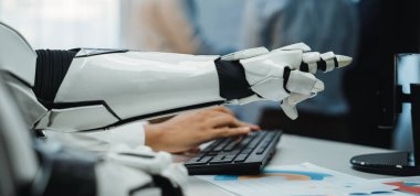 Çağdaş ofis ortamında bir klavyeye robotik bir kol yazarken detaylı bir bakış açısı, iş adamlarının bulanık figürleriyle birlikte teknoloji ve yenilik sergiliyor. Noogenesis