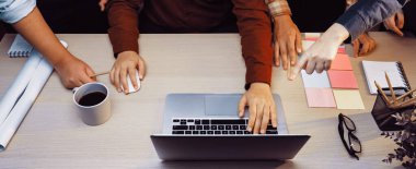 A dynamic office setting showcasing a collaborative team meeting, featuring hands engaged around a laptop, coffee, sticky notes, and bustling creative energy. SACTR