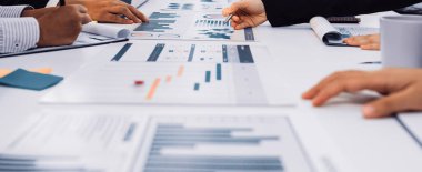 Bir grup iş adamı toplantı sırasında işbirliği yaparak çeşitli mali tablolar ve raporları analiz ederken, modern teknolojiyi şık bir ofis ortamında kullanıyorlar. SACTR